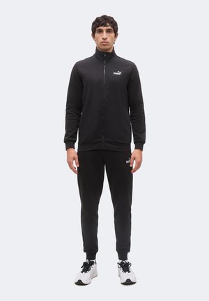 Mand står fremadvendt iført et sort Puma-joggingtøj og hvide sneakers på en ensfarvet lys baggrund.