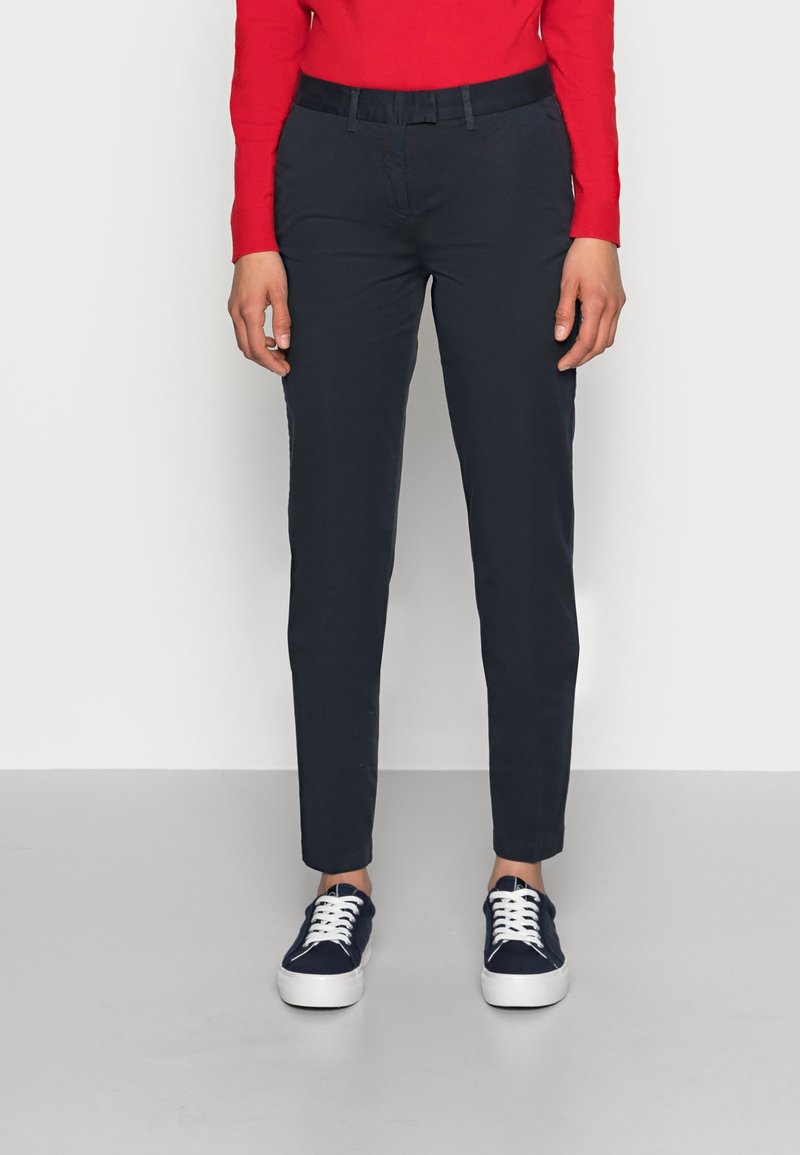 Personne portant un pantalon slim bleu marine foncé, des baskets bleu marine avec des lacets blancs et un haut à manches longues rouge, debout devant un fond uni.