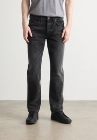 Jeans de mezclilla negra con un acabado desgastado, diseño de pierna recta, cinco bolsillos y costuras visibles. Llevados con zapatillas negras.