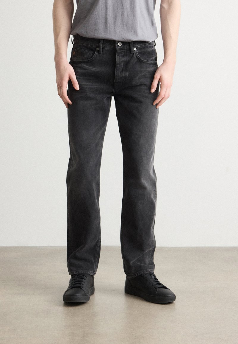 Jeans de mezclilla negra con un acabado desgastado, diseño de pierna recta, cinco bolsillos y costuras visibles. Llevados con zapatillas negras.