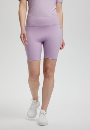 Mujer con pantalones cortos ajustados de ciclista color púrpura claro y zapatillas blancas, caminando hacia adelante sobre un fondo gris liso.