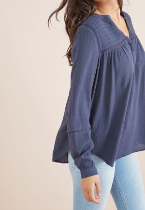 Woman wearing a navy blue long-sleeve textured blouse with lace details, paired with light blue denim jeans, against a beige background.