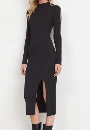 Vrouw in een zwarte geribbelde midi-jurk met lange mouwen, een voorste split en merktekst langs de mouwzomen.