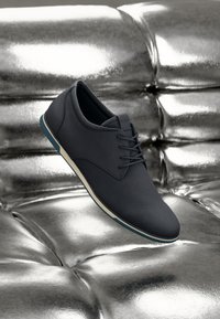 Mørkegrå sneakers med glatte læder- og tekstiloverflader, med blå accentsål og hvid mellemsål, vist på en metallisk overflade.