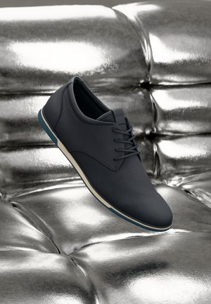 Dark gray sneaker with smooth leather and fabric textures, featuring blue accent sole and white midsole, displayed on a metallic surface.