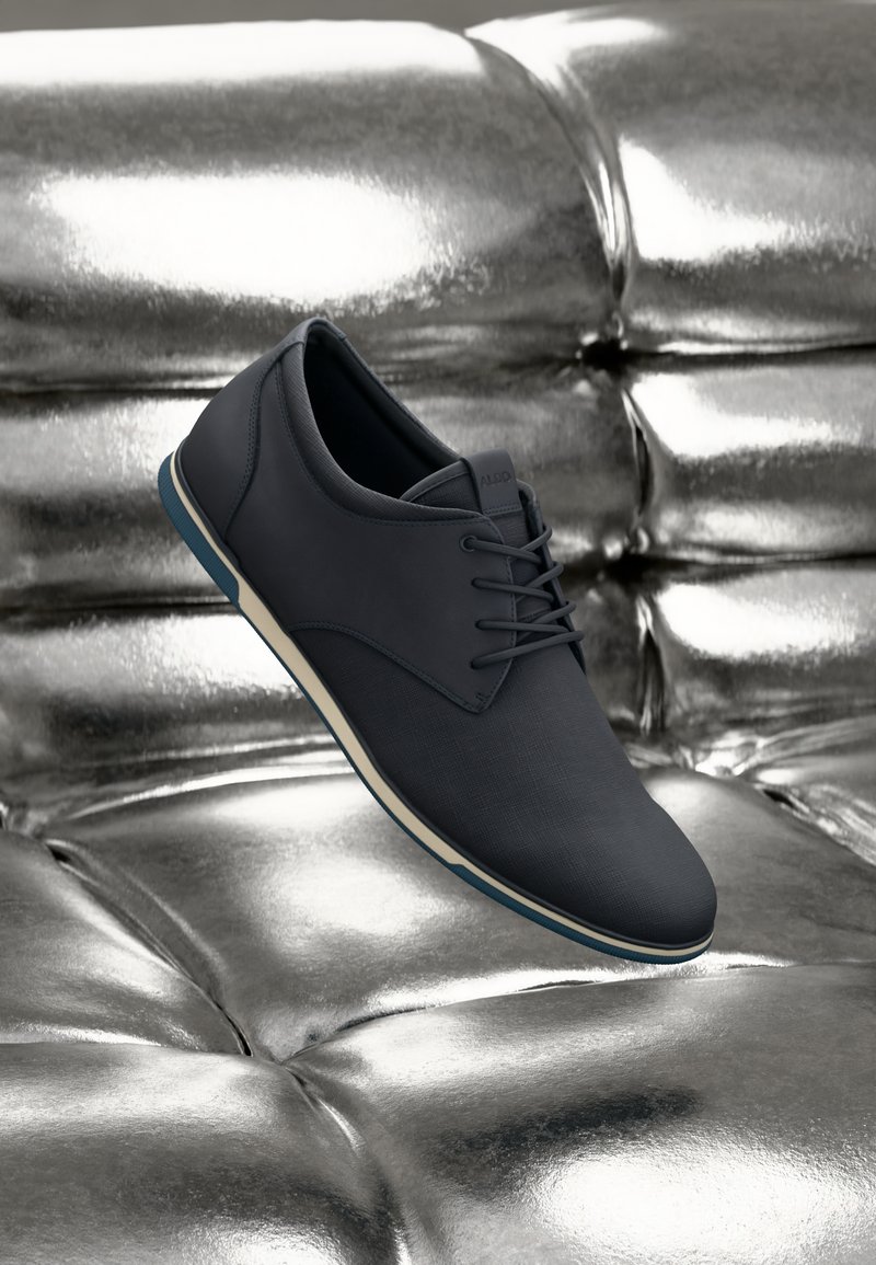 Mørkegrå sneakers med glatte læder- og tekstiloverflader, med blå accentsål og hvid mellemsål, vist på en metallisk overflade.