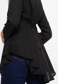 Haut noir à manches longues avec une taille cintrée, doté d'un ourlet froncé et de détails de boutons le long du dos, fabriqué en tissu léger.