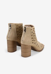 Botas de tobillo de ante beige con detalles recortados, tacón cuadrado y cierre de cremallera. Presenta un tacón texturizado y estriado, además de un diseño de punta afilada.