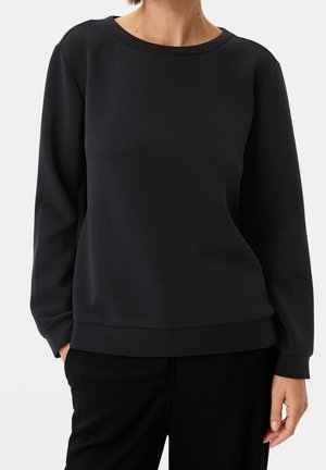 Personne portant un sweat-shirt noir uni à manches longues et col rond, les mains partiellement dans les poches d'un pantalon noir.