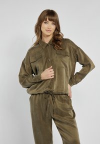 Zielonooliwkowy dres z materiału kordurowego, z koszulą zapinaną na guziki, dwiema kieszeniami na piersi i elastycznym paskiem. Miękka tekstura i luźny krój.