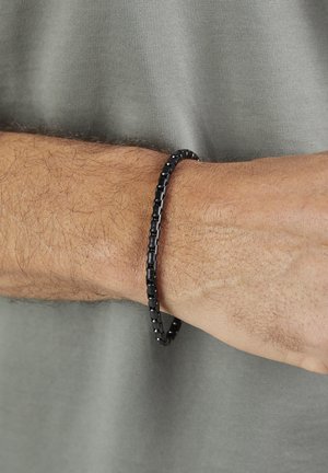 Sort perlearmbånd båret på et behåret håndled med lys brun hud mod en lys grå tekstureret stofbaggrund.