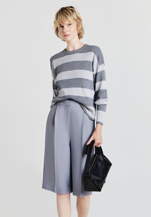 Maglione a righe grigio e bianco in maglia abbinato a pantaloni palazzo grigio chiaro. Il modello tiene una piccola borsa in pelle nera, presenta una vestibilità rilassata e una texture a coste.
