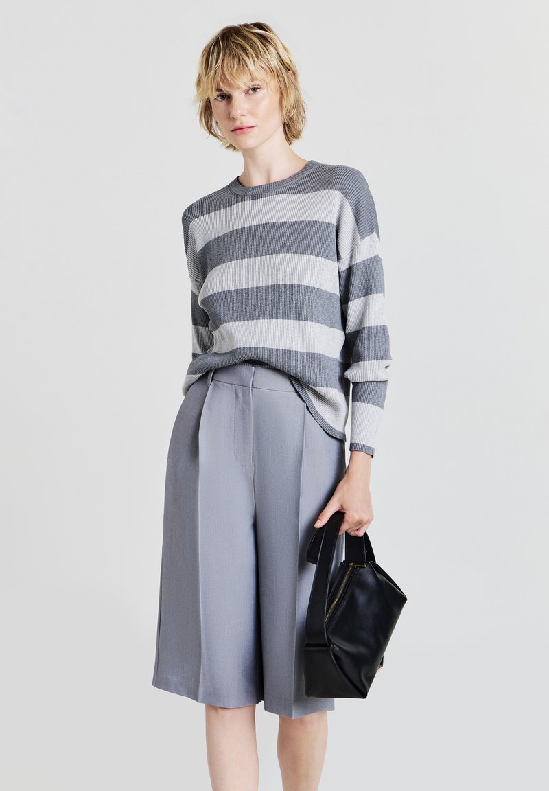 Maglione a righe grigio e bianco in maglia abbinato a pantaloni palazzo grigio chiaro. Il modello tiene una piccola borsa in pelle nera, presenta una vestibilità rilassata e una texture a coste.