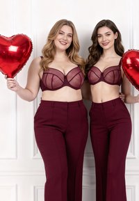 Dvě usměvavé ženy držící červené balóny ve tvaru srdce, oblečené ve stejných burgundských kalhotách a průsvitných podprsenkách, stojící před bílou zdí s panely.