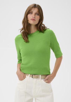 Pull vert en tricot avec texture côtelée, col rond et manches courtes, associé à un pantalon blanc taille haute et une ceinture beige.