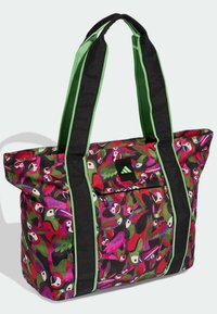 Bolsa tote con asas negras y verdes, estampado de loro multicolor en rojo, rosa, verde y morado, y bolsillo frontal con cremallera y parche con el logo.