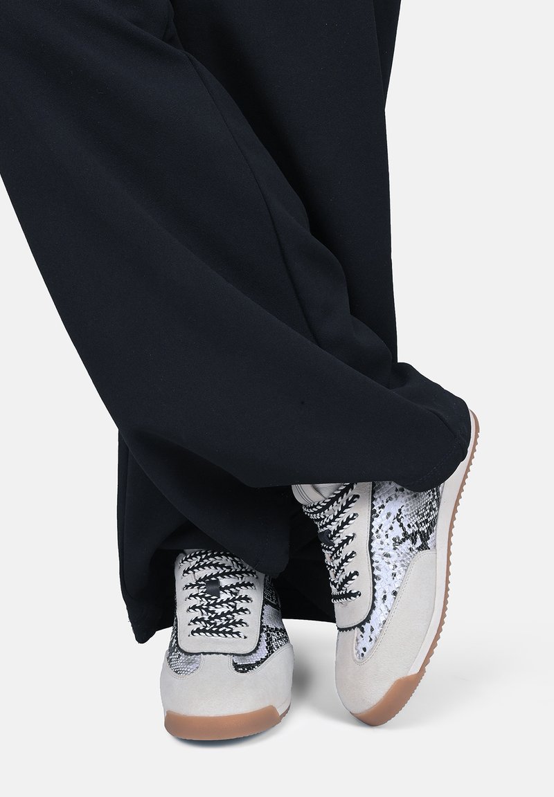 Osoba ubrana w czarne spodnie i białe trampki z czarnym wzorem węża oraz beżowymi akcentami, stojąca ze skrzyżowanymi stopami.