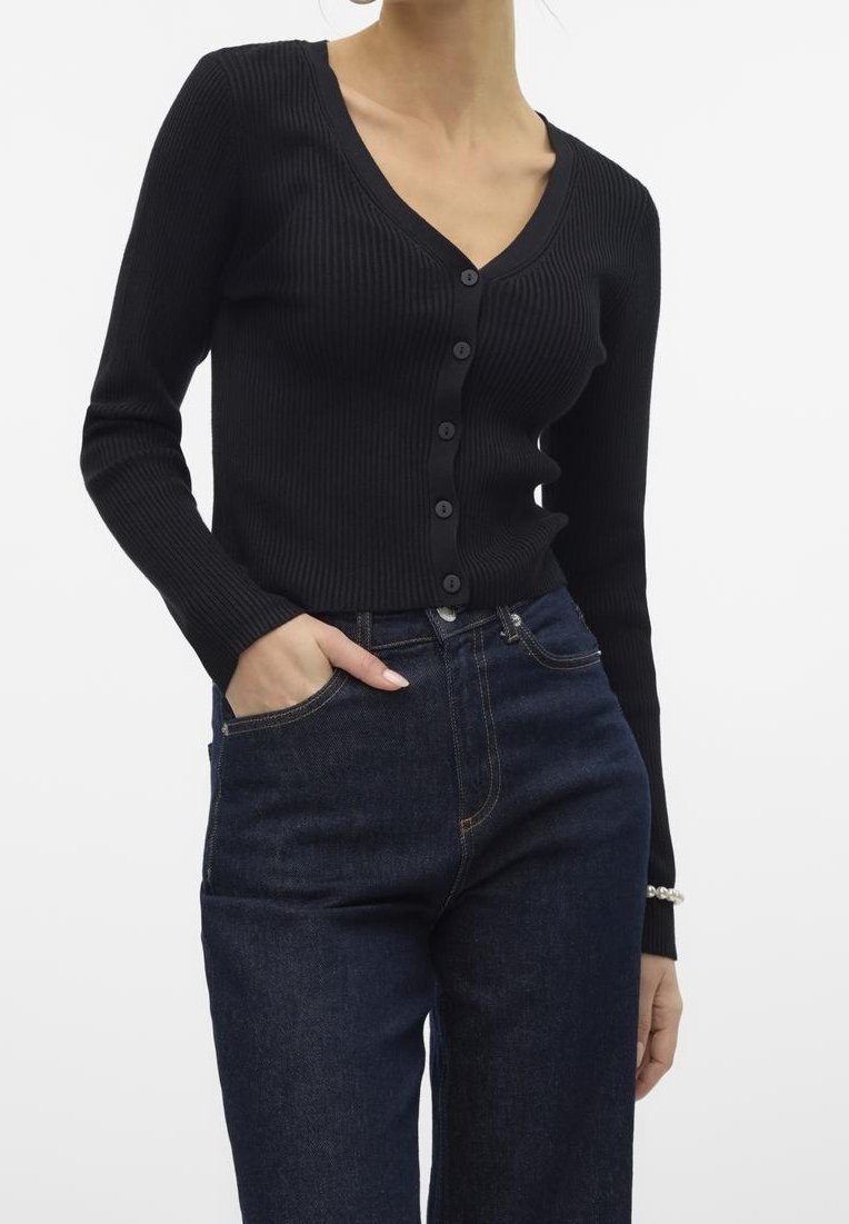 Femme portant un cardigan noir côtelé à boutons avec manches longues et un jean taille haute bleu foncé, une main dans la poche, sur fond blanc.