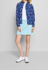 Bunda s modrým paisley vzorem, bílý polo tričko a světle modrá sukně; model má na sobě bílé sportovní boty. Měkké materiály, volný střih a viditelný zip.