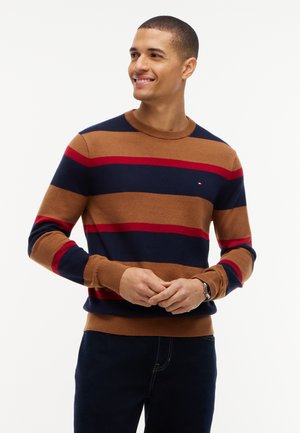 Mand smilende, iført en brun, marineblå og rødstribet sweater og mørke bukser, med hænderne løst foldet foran.