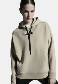 Béžová mikina s kapucí z měkkého materiálu, s černou šňůrkou, přední kapsou a žebrovanými manžetami. Minimalistický design s čistými liniemi.