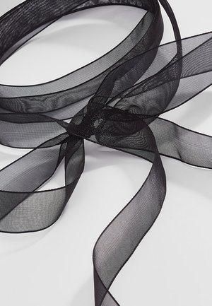 Musta läpikuultava organzalentti, löysästi kierrettynä ja levitettynä valkoiselle pinnalle, paljastaen sen läpinäkyvän ja kevyen rakenteen.