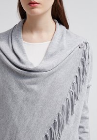 Grijze gebreide omslag met een brede col, voorzien van een decoratieve knoop en franje randen. Zachte textuur, valt losjes over de schouders.