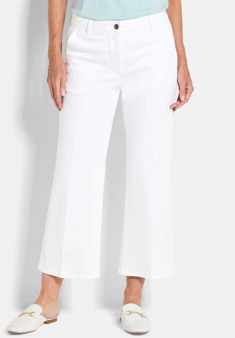 Vrouw in witte cropped wijde broek, lichtblauw shirt en witte instap loafers met gouden gespdetail.