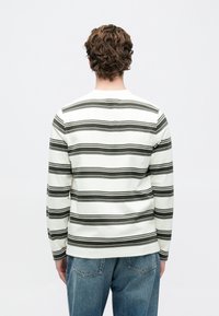 Άνδρας με καστανά σγουρά μαλλιά, φορώντας λευκό πουκάμισο με μακριά μανίκια και οριζόντιες μαύρες ρίγες, καθώς και μπλέ τζιν, γυρισμένος πλάτη σε μια απλή φόντο.