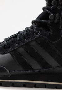 Botte de randonnée noire composée d'un mélange de cuir et de daim. Dispose de lacets visibles, d'œillets métalliques et du design signature à trois bandes.