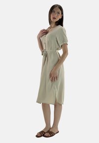 Abito beige lungo fino al ginocchio con maniche a puff corte, bottoni frontali, cintura in vita e spacco laterale. Tessuto morbido con finitura texturizzata. Sandali piatti marroni.