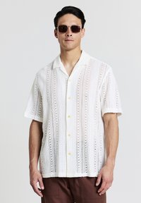 Chemise blanche à manches courtes présentant des motifs en dentelle, un col et des boutons jaunes. Portée avec un pantalon marron et des lunettes de soleil rectangulaires.