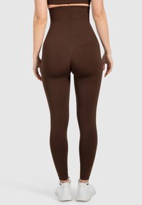 Hoge taille bruine legging gemaakt van rekbaar materiaal, met een gladde textuur en een aansluitende pasvorm, gedragen met witte sportschoenen.