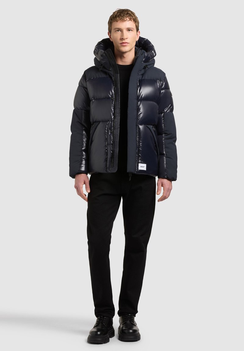 Homme portant une veste matelassée bleu foncé brillante avec capuche, pantalon noir et chaussures noires, debout devant un fond clair uni.