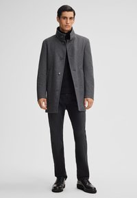 Grau melierter Wollmischmantel mit hohem Kragen, drei Knöpfen und seitlichen Taschen. Kombiniert mit schwarzen Jeans und schwarzen Lederschuhen.
