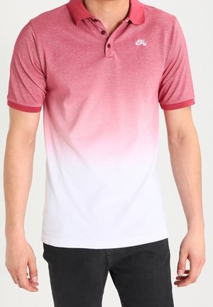 Mand iført en kortærmet rød til hvid gradient polo med Nike SB-logo og sorte bukser, stående med arme afslappet.
