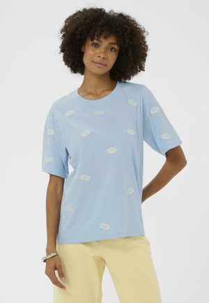 Młoda kobieta o kręconych włosach, ubrana w jasnoniebieską koszulkę z białymi nadrukami w kształcie ust oraz jasnopalone spodnie, stojąca z jedną ręką na biodrze.