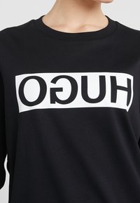 Persona con una camiseta negra que tiene un rectángulo blanco con la palabra "HUGO" impresa en letras negras, invertida horizontalmente.