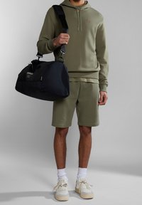 Olivově zelená mikina s kapucí a kraťasy z měkké tkaniny; nese černou tašku s logem. Má na sobě bílé tenisky a ponožky do poloviny lýtek.