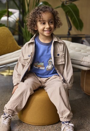 Child with curly hair wearing a blue shirt and beige jacket, sitting on a yellow round cushion indoors with plants in the background.
