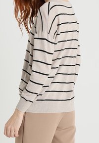 Femme portant un pull beige à manches longues avec des rayures horizontales noires et un pantalon beige, vue de dos et de côté sur un fond clair.