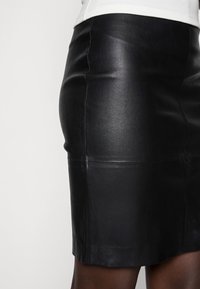Falda de cuero negra con un diseño ajustado, que presenta una textura suave y costuras visibles a lo largo de las uniones. El dobladillo termina por encima de la rodilla.
