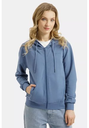 Kobieta o blond włosach, ubrana w niebieską bluzę na zamek z kapturem, na białej koszuli i jasnych niebieskich dżinsach, pozująca z jedną ręką w kieszeni.