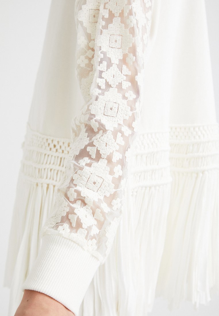 Garbato bianco con un corpo solido e maniche in pizzo semi-trasparente con motivi geometrici, rifinito con frange all'orlo e polsini testurizzati.