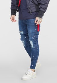 Vaqueros ajustados de mezclilla azul con desgastes rotos y detalles de salpicaduras de pintura. Combinados con zapatillas blancas y una chaqueta bomber oscura.