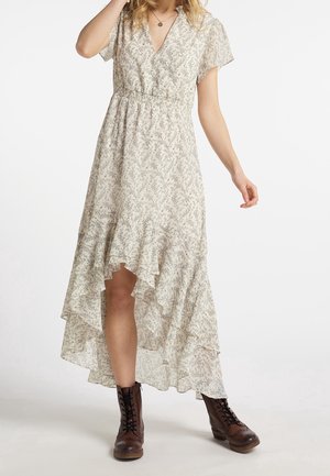 Frau in einem hellen, floralen High-Low-Kleid, kombiniert mit dunkelbraunen Schnürstiefeln, posiert mit einem erhobenen Arm in der Nähe ihres Gesichts.