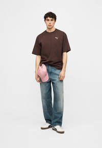 Νεαρός άνδρας στέκεται φορώντας καφέ μπλουζάκι Puma, φαρδιά μπλε τζιν, λευκά αθλητικά παπούτσια, κρατώντας ροζ καπέλο Puma στο αριστερό χέρι, με φόντο απλό.