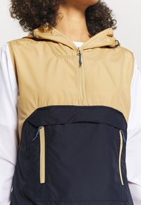 Lehká dvoubarevná vesta se zlatou horní částí a tmavě modrým spodním panelem, vybavená předním zipem, dvěma kapsami na zip a kapucí.