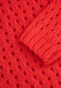 Czerwony sweter z dzianiny z wzorem ażurowym, teksturowaną włóczką i ściągaczem. Materiał ma lekko puszysty wygląd.