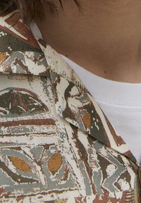 Gros plan sur une personne portant une chemise à motifs avec des formes abstraites marron, blanches et grises sur un t-shirt blanc à col rond.